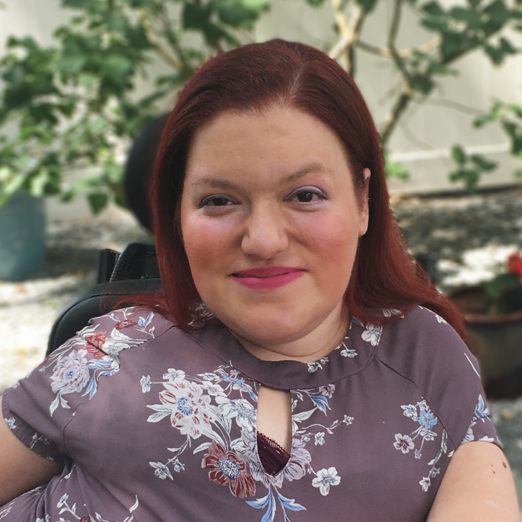 A white woman with long auburn hair, wearing a purple floral top and fuchsia lipstick. She is smiling at the viewer.