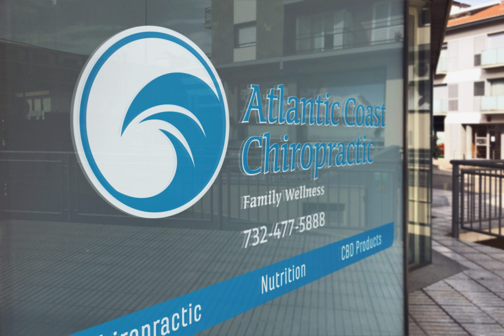 The Atlantic Coast Chiropractic logo is printed on a plate glass window above business details, phone number, and a blue horizontal bar printed with services offered. The logo features a blue stylized wave inside a circle to the leff of the words 'Atlantic Coast Chiropractic' (also blue).