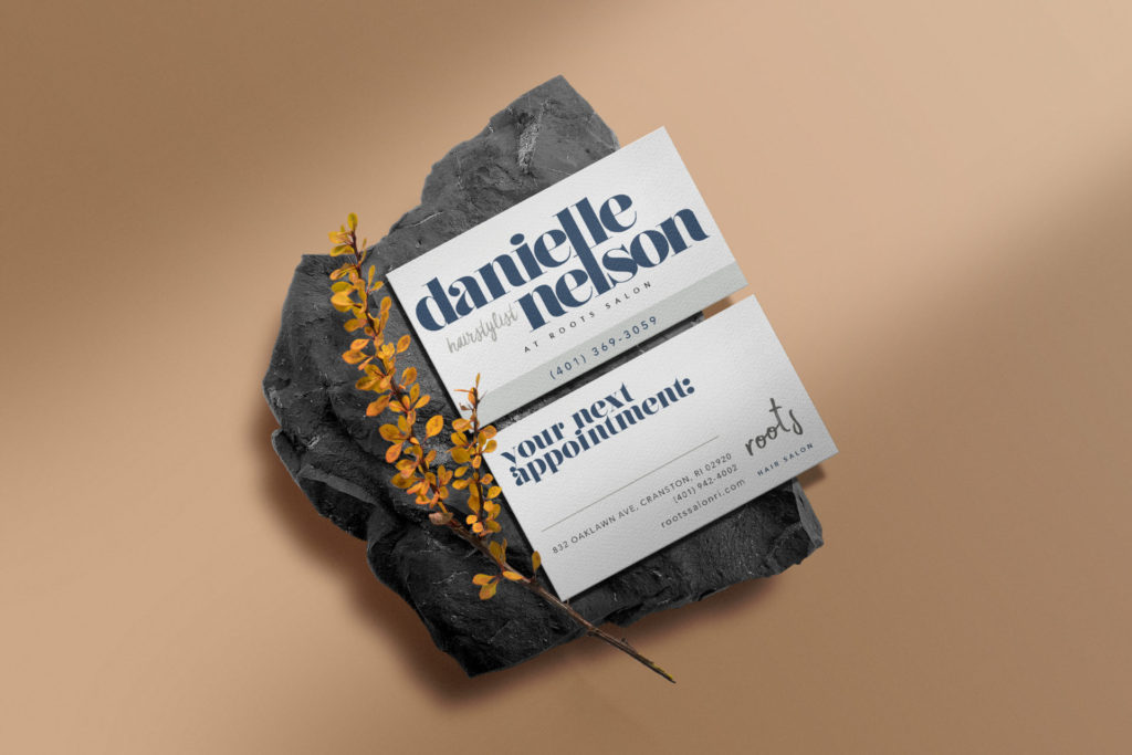two white cards are printed with the front and back side of the business card for danielle nelson. The cards and placed diagonally on a dark gray rock beside a spring of yellow-orange dried foliage. Everything is placed on a tan background with stylish shadows cast over the top left and bottom right of the image.