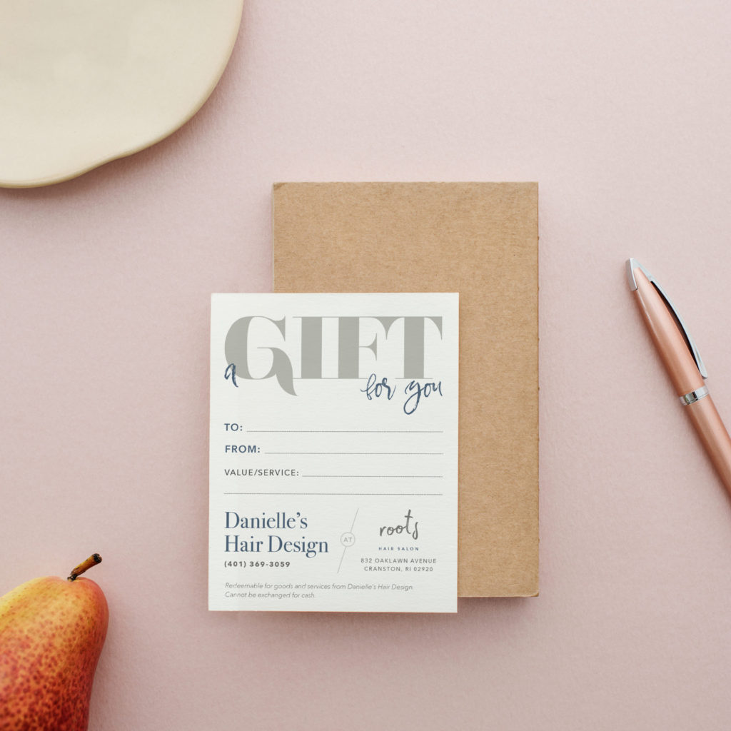 a vertical white gift certificate is placed on a tan wood block on a light pink background. A rose gold pen, red pear, and white tray are arranged around the periphery of the image.