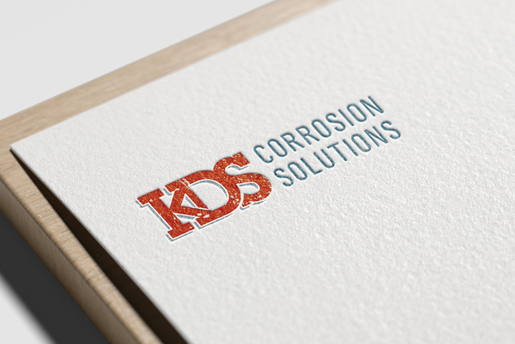 A white sheet of thick, textured paper sits on a wooden platform on a light gray background. The KDS Corrosion Solutions logo is imprinted onto the white paper.