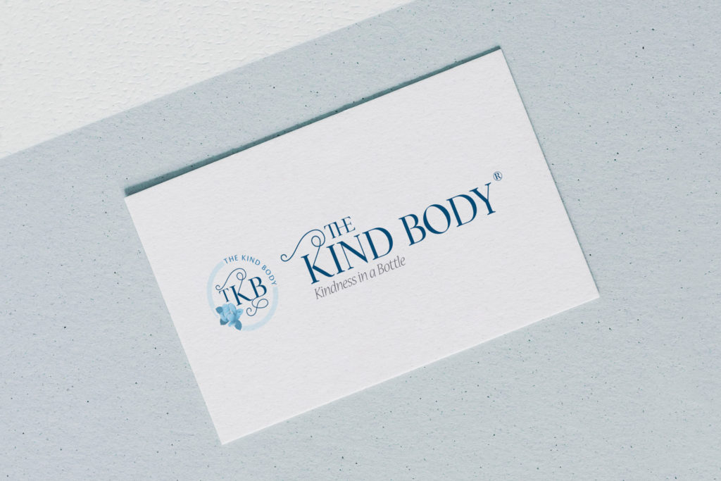 a white card sits on top of a soft blue paper diagonally on a white background. The card is printed with the detailed version of The Kind Body logo