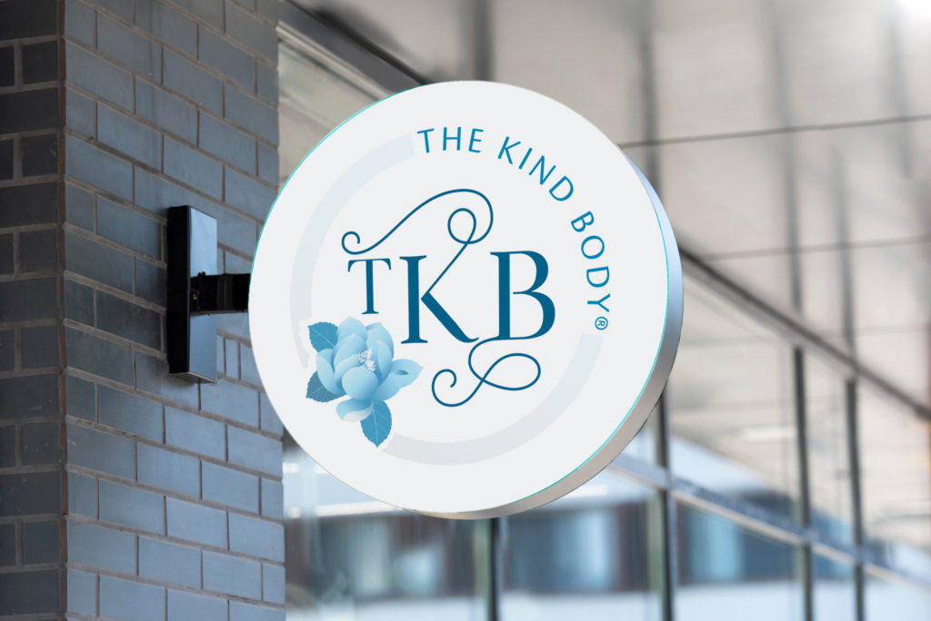 A round white storefront sign is printed with the initials TKB surrounded by a blue flower, pale blue circle border, and 'The Kind Body'